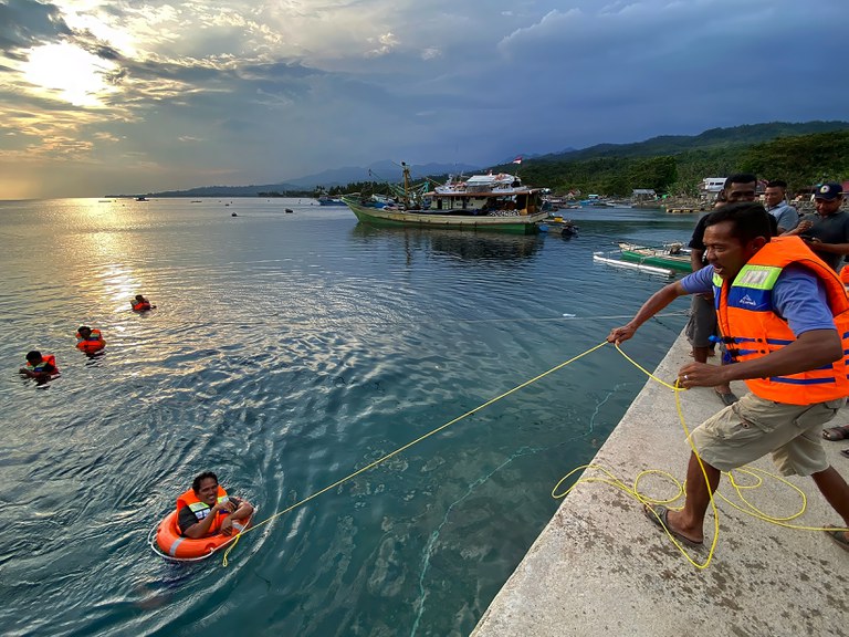 Fishermen practice a safety drill in the open water, with one man standing on concrete dock throwing a bright orange life raft.