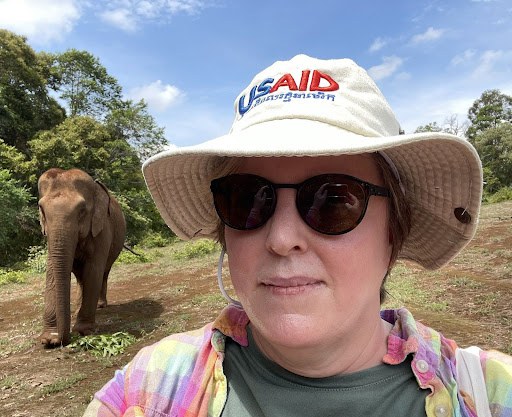 A woman in sunglasses and a hat smiles with an elephant in the background.