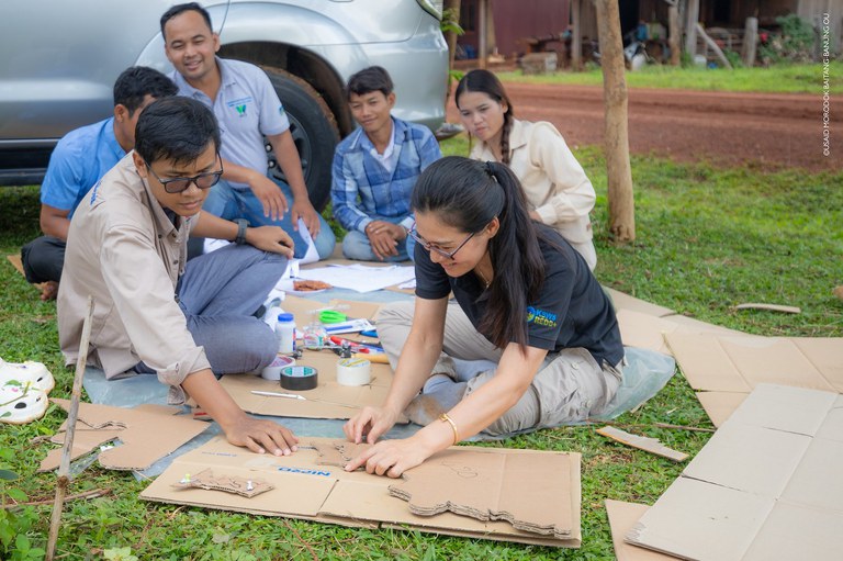 A group of people sit in grass and glue pieces of a cardboard map together